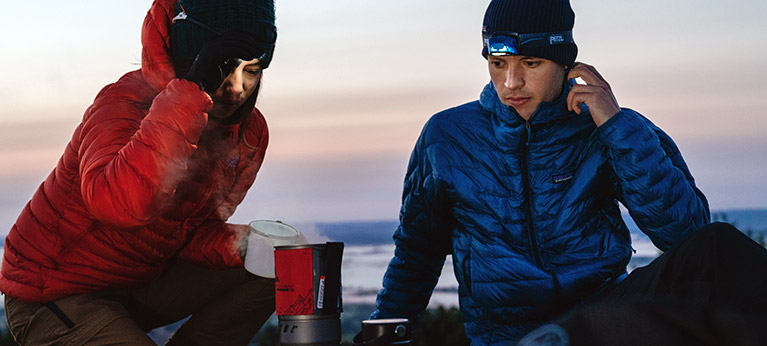 A man and woman sitting over a camping stove wearing insulated jackets